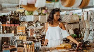 Eine junge Frau in einem weißen Kleid steht an einem exotischen Basar und sieht sich die verschiedenen Waren an.