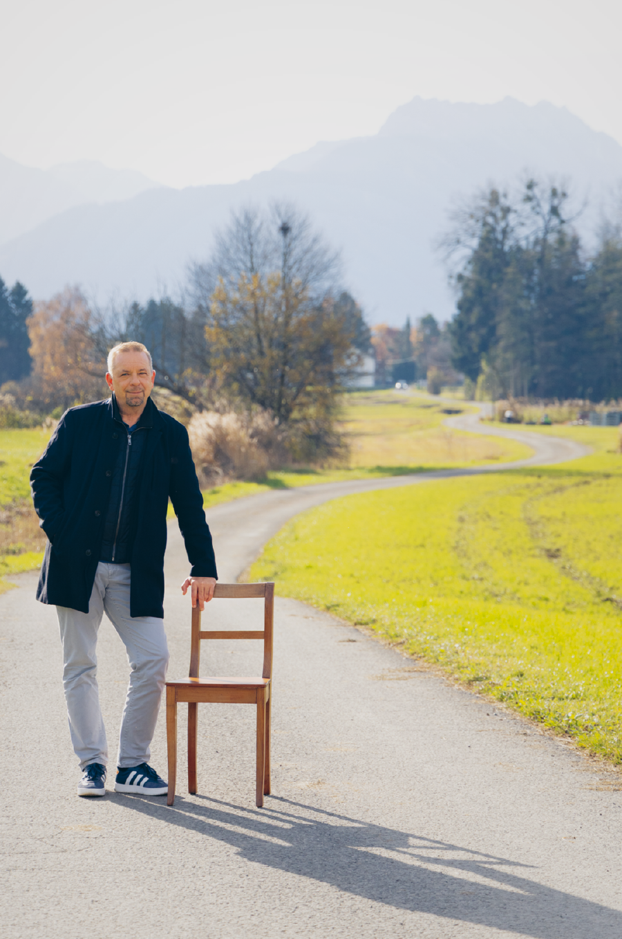 Oliver Loacker steht auf einer Stra&szlig;e und st&uuml;tzt sich auf einen Stuhl. Nebenan sieht man eine gr&uuml;ne Wiese, im Hintergrund ein Bergpanorama.