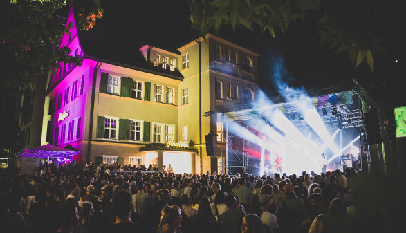 N&auml;chtliches Open-Air-Konzert beim Schaffarei Festival mit einer dicht gedr&auml;ngten Menschenmenge vor einer beleuchteten B&uuml;hne und einem violett-gelb angestrahlten historischen Geb&auml;ude.