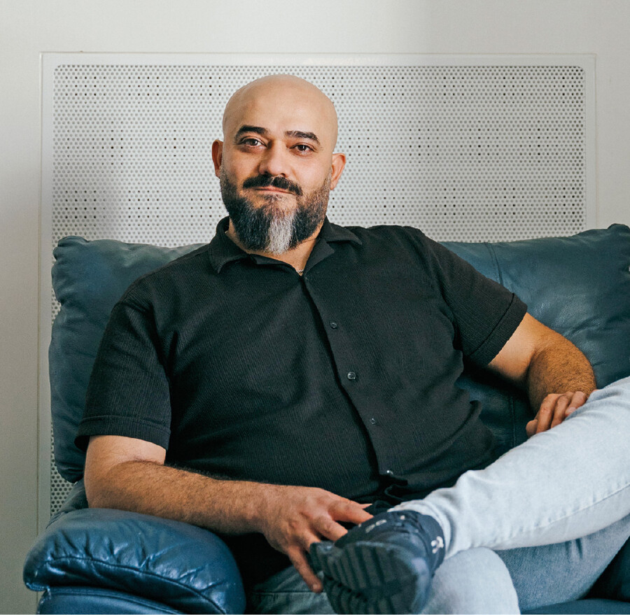 Portr&auml;t eines glatzk&ouml;pfigen Mannes mit dunklem Vollbart und ergrauter Kinnpartie. Er tr&auml;gt ein schwarzes Kurzarmhemd sowie eine helle Jeans und sitzt entspannt mit &uuml;berschlagenen Beinen in einem dunklen Ledersessel. Im Hintergrund ist eine wei&szlig;e Wand mit einem gelochten L&uuml;ftungsgitter zu sehen.