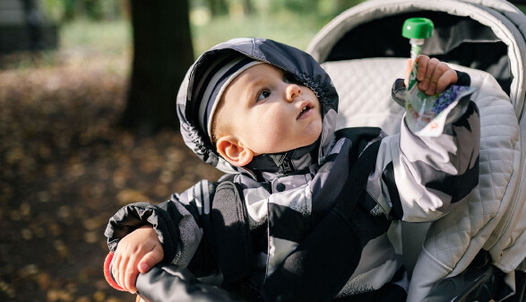 Ein Baby sitzt angeschnallt in einem Kinderwagen und h&auml;lt eine Trinkflasche in der Hand. Der Kinderwagen steht drau&szlig;en in einer gr&uuml;nen Umgebung.