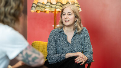 Eugenia Stamboliev sitzt im Gespr&auml;ch auf einem gelben Sessel vor einer roten Wand und spricht engagiert in Richtung ihres Gegen&uuml;bers.