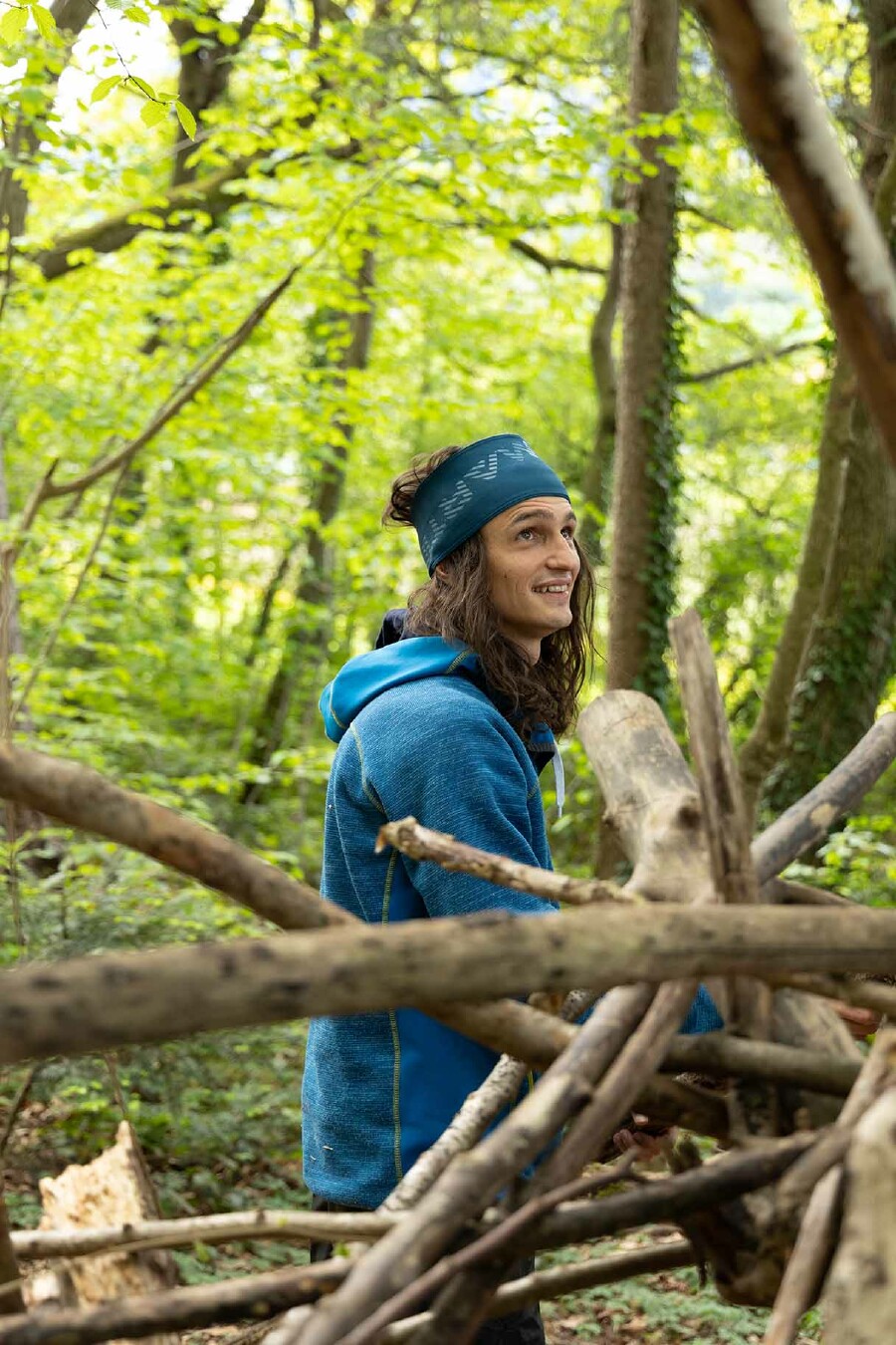 Ein Mann mit langen, braunen Haaren in einer blauen Jacke, Simon Engstler, steht lächelnd in einem Wald.