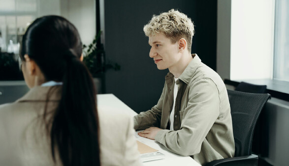 Ein junger, blonder Mann in einem Hemd sitzt in einer Gesprächssituation mit einer dunkelhaarigen Frau in einem Blazer an einem Besprechungstisch und lächelt selbstsicher.