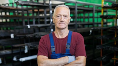 &Auml;lterer Arbeiter in Latzhose steht mit verschr&auml;nkten Armen in einem Industrielager und blickt ernst in die Kamera.