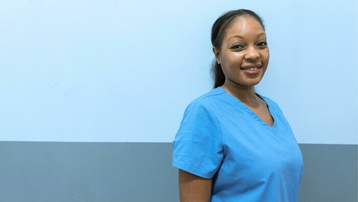 L&auml;chelnde Pflegekraft in blauer medizinischer Arbeitskleidung vor hellblauem Hintergrund &ndash; symbolisiert das Pflegepersonal, das von der neuen Schwerarbeitsregelung profitieren kann.