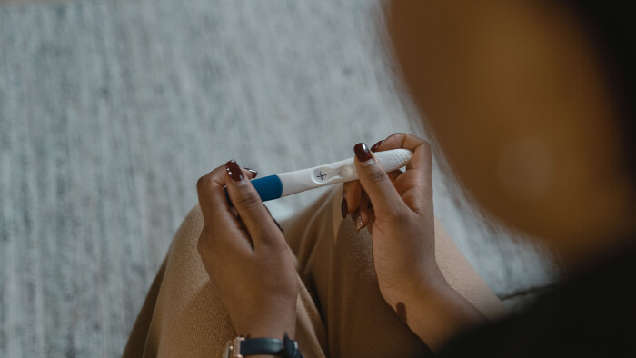 Nahaufnahme aus der Perspektive &uuml;ber die Schulter einer sitzenden Person, die einen positiven Schwangerschaftstest mit zwei H&auml;nden h&auml;lt. Das Kontrollfenster des wei&szlig;en Tests mit blauer Kappe zeigt deutlich ein Pluszeichen. Die Person tr&auml;gt eine Armbanduhr mit dunklem Band und sitzt auf einem hellen Teppichboden