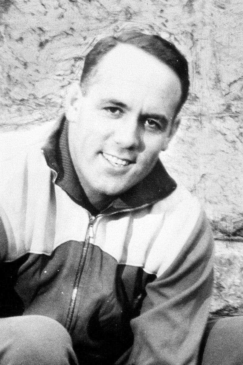 Historische Schwarz-Wei&szlig;-Fotografie eines jungen Mannes mit dunklen, kurzen Haaren, der direkt in die Kamera l&auml;chelt. Er tr&auml;gt eine sportliche Trainingsjacke mit Rei&szlig;verschluss und hohem Kragen. Im Hintergrund ist eine helle, strukturierte Steinmauer zu sehen.