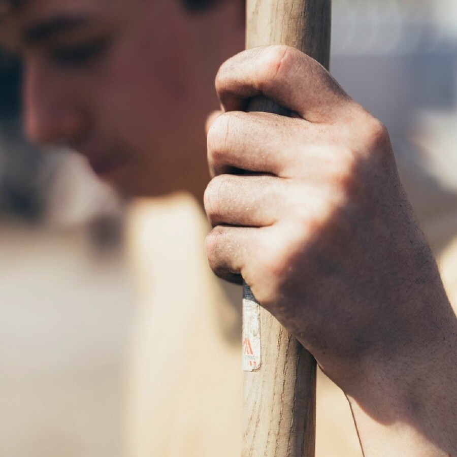 Nahaufnahme einer Hand mit Erde, die fest ein Holzwerkzeug greift – Symbol für körperliche Arbeit und praktische Ausbildung im Gartenbau. Im Hintergrund unscharf das Gesicht des Lehrlings, umgeben von natürlichem Licht. Authentisches Bild für handwerkliches Können, Outdoor-Berufe und nachhaltige Gartenpflege.
