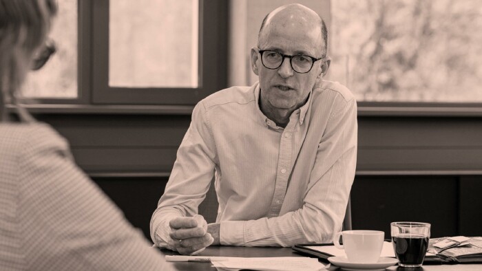 Schwarz-Wei&szlig;-Aufnahme von Thomas Melmer w&auml;hrend eines Interviews. Er sitzt an einem Tisch mit Kaffeetasse und Unterlagen und spricht konzentriert mit einer Person im Vordergrund, die nur von hinten zu sehen ist.