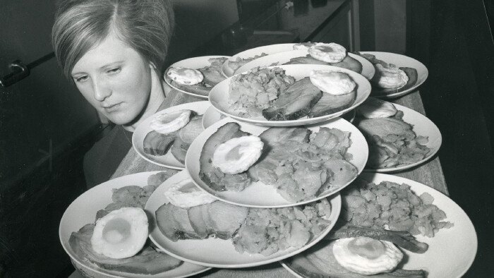 Schwarz-wei&szlig;-Bild einer Kellnerin 1967. Sie tr&auml;gt auf der Schulter ein gro&szlig;es Tablett voller beladener Teller.