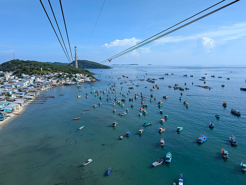 Der asiatische Raum – wie hier Vietnam – gefällt Seilbahntechniker Elmar Röck besonders. © Elmar Röck Dutzende kleine, bunte Fischerboote liegen vor einem Sandstrand im blauen Meer. Im Hintergrund sieht man grün bewachsene Inseln und dazwischen eine riesige Seilbahnstütze.