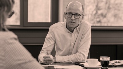 Schwarz-Wei&szlig;-Aufnahme von Thomas Melmer w&auml;hrend eines Interviews. Er sitzt an einem Tisch mit Kaffeetasse und Unterlagen und spricht konzentriert mit einer Person im Vordergrund, die nur von hinten zu sehen ist.