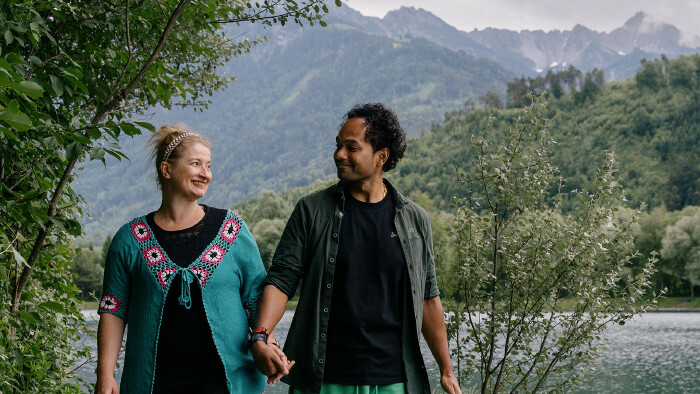 Ein l&auml;chelndes Paar h&auml;lt H&auml;ndchen und blickt sich liebevoll an, w&auml;hrend es in einer alpinen Landschaft spazieren geht. Links steht eine Frau mit hochgesteckten blonden Haaren, einem Haarband und einer markanten t&uuml;rkisfarbenen Strickjacke mit rosa-wei&szlig;en H&auml;kelmustern. Rechts steht ein Mann mit dunklen, lockigen Haaren, einem schwarzen T-Shirt und einem offenen, dunkelgr&uuml;nen Hemd. Im Hintergrund erstreckt sich ein ruhiger See, hinter dem dicht bewaldete Bergh&auml;nge und schroffe, wolkenverhangene Gebirgsz&uuml;ge aufragen. Am linken und rechten Bildrand rahmen gr&uuml;ne Str&auml;ucher und B&auml;ume die Szene ein.