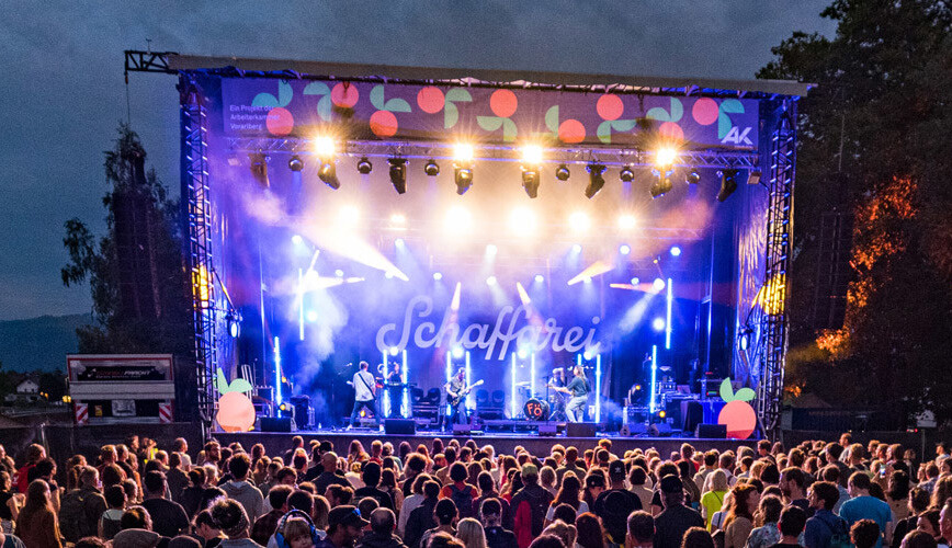 Abendliches Freiluftkonzert auf dem Schaffarei Festival: Eine Band steht auf einer beleuchteten Festivalb&uuml;hne unter hellen Scheinwerfern und Nebeneffekten. Im Hintergrund der B&uuml;hne ist der geschwungene Schriftzug &bdquo;Schaffarei&ldquo; zu sehen. Im Vordergrund steht ein dicht gedr&auml;ngtes Publikum von hinten gesehen und blickt zur B&uuml;hne.