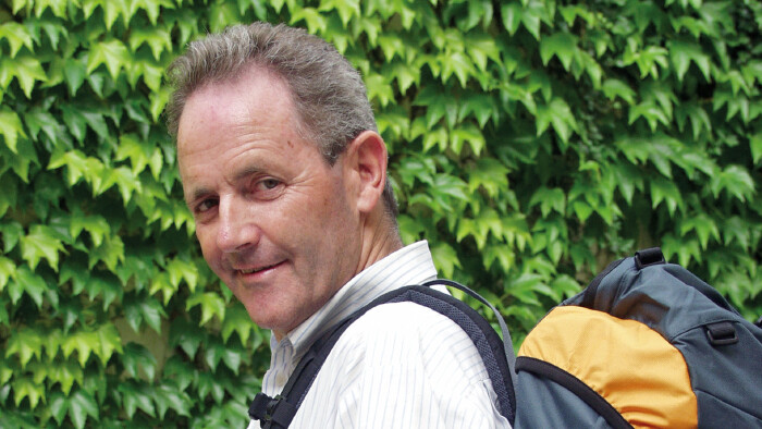 Alt-AK-Pr&auml;sident Josef Fink dreht sich l&auml;chelnd zur Kamera um, einen Rucksack auf dem R&uuml;cken. Der Rucksack steht symbolisch f&uuml;r das von ihm mitinitiierte Abfertigungsmodell &sbquo;Abfertigung neu', das Arbeitnehmerinnen und Arbeitnehmer wie einen mitwachsenden Rucksack durchs Berufsleben begleitet. Im Hintergrund eine gr&uuml;ne Blattwand.