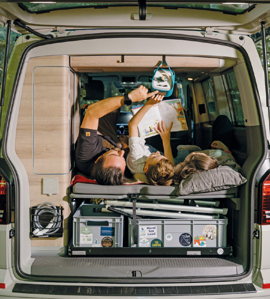Blick durch die ge&ouml;ffnete Heckklappe eines ausgebauten Camper-Vans. Auf der Liegefl&auml;che im Innenraum liegen drei Personen: Ein Mann bedient eine oben aufgeh&auml;ngte Toniebox, ein Kind liest in einem Kinderbuch und eine weitere Person liegt daneben. Unter der Liegefl&auml;che befinden sich Kisten mit Camping-Stickern und Verstauraum.