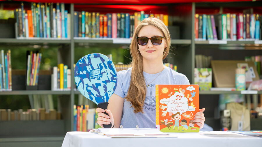 Eine Frau mit hellbraunem Haar und Sonnenbrille steht hinter einem Tisch mit wei&szlig;er Tischdecke im Freien. Hinter ihr befindet sich ein gro&szlig;er, mobiler B&uuml;cherregalwagen aus Metall mit einem roten Dach, der reichlich mit B&uuml;chern gef&uuml;llt ist. Sie l&auml;chelt leicht und h&auml;lt in ihrer rechten Hand ein buntes Kinderbuch mit dem Titel &bdquo;50 Reime f&uuml;r Kleine&ldquo; und in ihrer linken Hand einen runden, blau gemusterten F&auml;cher. Sie tr&auml;gt ein graues T-Shirt mit einem Print. Der Hintergrund zeigt gr&uuml;ne Pflanzen und ein Holzgeb&auml;ude.