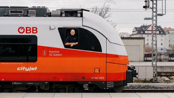 Lokf&uuml;hrer Holger Hansen lehnt aus dem Seitenfenster eines rot-wei&szlig;en &Ouml;BB-Cityjet-Zuges am Bahnsteig und blickt in die Kamera; im Hintergrund sind Gleise, ein Signal und Wohnh&auml;user zu sehen.