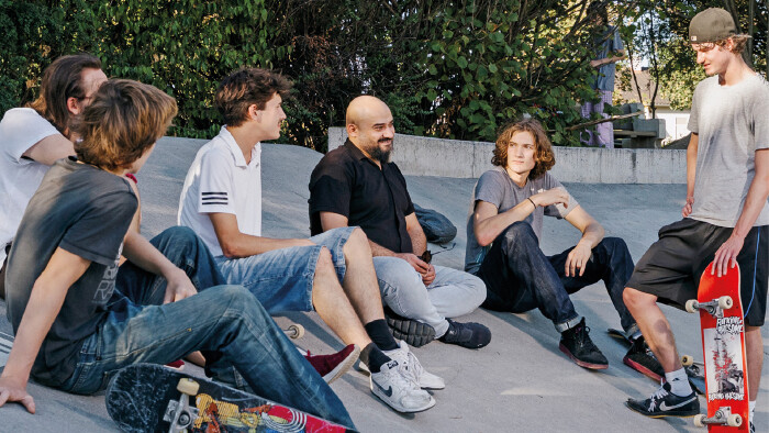 Eine Gruppe von jungen M&auml;nnern sitzt und steht entspannt in einem Skatepark aus Beton und unterh&auml;lt sich. Im Vordergrund liegen und stehen Skateboard-Decks, rechts h&auml;lt ein junger Mann mit Basecap ein rotes Skateboard fest. Im Hintergrund sind gr&uuml;ne B&auml;ume und Str&auml;ucher zu sehen.