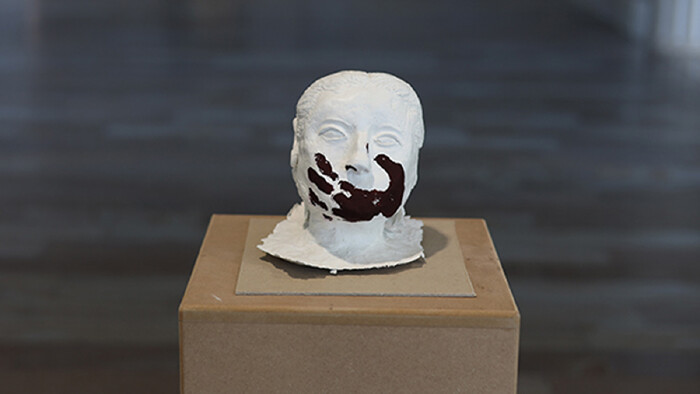 Eine Plastik eines Kopfes steht auf einem Sockel, mit schwarzer Farbe ist ein Handabdruck über dem Mund zu sehen.