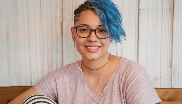 Eine Frau mit blauen Haaren und einer Brille sitzt auf einer Bank und blickt in die Kamera.