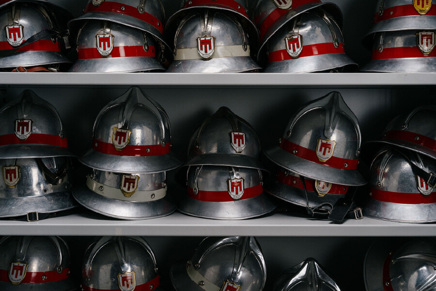 Eine frontale Nahaufnahme von mehreren Reihen alter, silberner Feuerwehrhelme aus Metall, die ordentlich in einem grauen Regal nebeneinander und &uuml;bereinandergestapelt aufgereiht sind. Die Helme im traditionellen Design besitzen einen markanten roten oder wei&szlig;en Querstreifen auf der Vorderseite. Mittig auf der Stirnseite jedes Helms ist ein wappenf&ouml;rmiges Emblem angebracht, das das Stadtwappen von Feldkirch (eine rote Kirchenfahne auf wei&szlig;em Grund) zeigt und mit dem darunter liegenden Schriftzug &bdquo;Tosters&ldquo; (Ortsfeuerwehr Tosters) versehen ist.