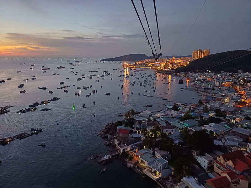 Bei so einer Aussicht – hier auf eine Bucht in Vietnam – macht auch eine Spätschicht nichts aus. © Elmar Röck Eine bebaute Bucht am Meeresrand bei Sonnenuntergang. In den Gebäuden und Straßen brennen die ersten Lichter, auf dem Meer treiben Fischerboote, die Sonne geht rot am Horizont unter.