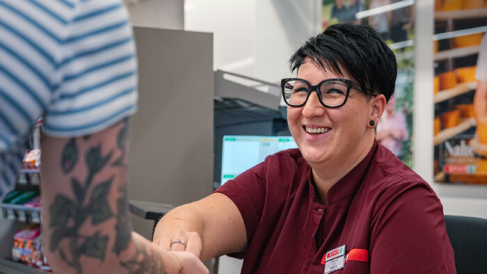 Eine l&auml;chelnde Kassiererin mit Brille, kurzen dunklen Haaren und einem weinroten SPAR-Arbeitshemd sitzt an einer Kasse. Sie reicht einer anderen Person, deren t&auml;towierter Arm und gestreiftes Oberteil im Vordergrund zu sehen sind, Wechselgeld oder ein Objekt.