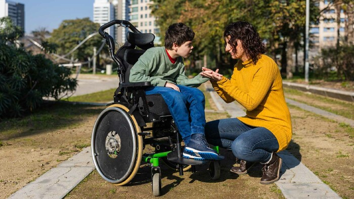Eine Frau kniet vor einem Jungen im Rollstuhl und hält seine Hand. Im Hintergrund ist ein Park zu sehen, die Sonne scheint.