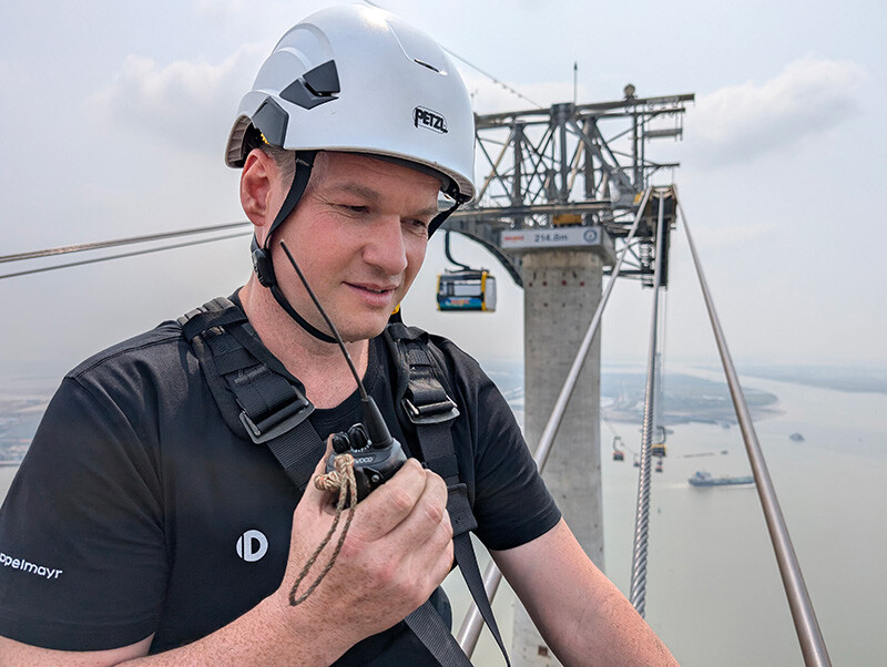 Elmar Röck ist seit zehn Jahren auf den Seilbahnstützen im Einsatz – zuletzt auf der höchsten Seilbahnstütze der Welt in Vietnam. © Elmar Röck Seilbahntechniker Elmar Röck spricht in ein Funkgerät, im Hintergrund sieht man eine Seilbahnstütze, auf der 214,8 Meter steht. Darunter sind nur Meer und zwei Inseln zu sehen.