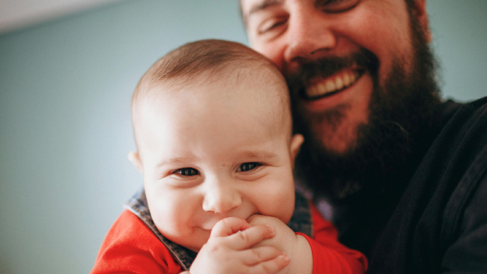 Ein lachendes Baby im roten Oberteil h&auml;lt die Hand im Mund, w&auml;hrend ein l&auml;chelnder Mann mit dunklem Bart seinen Kopf liebevoll an das Baby schmiegt.