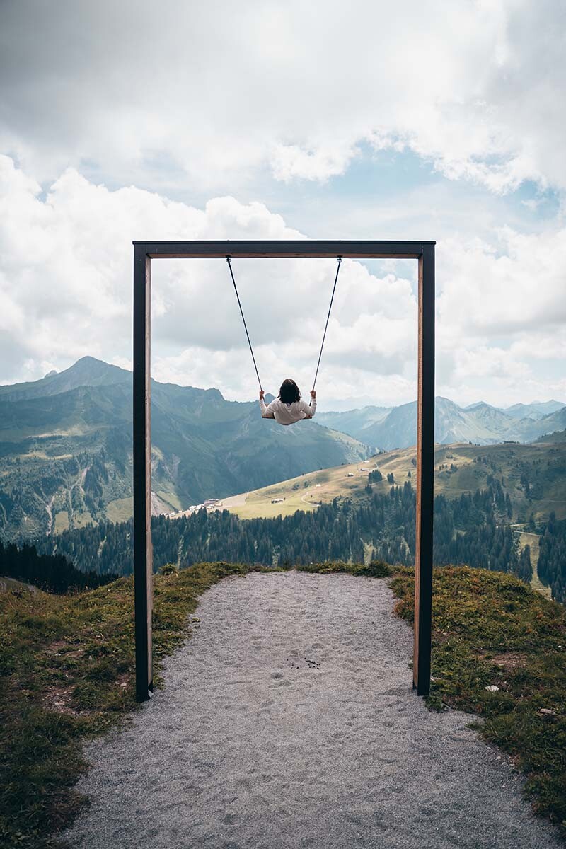 Eine Frau schaukelt auf einer großen Holzschaukel, im Hintergrund sind man ein Bergpanorama mit vielen Gipfeln.