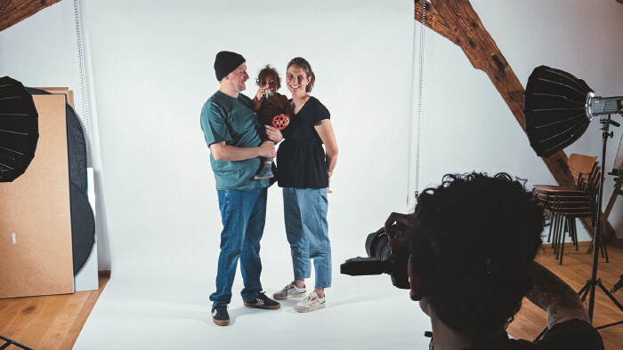 Ein Mann steht in einem Fotostudio und lacht das Kleinklind, das er auf dem Arm hält, an. Eine schwangere Frau steht daneben und lächelt in die Kamera.