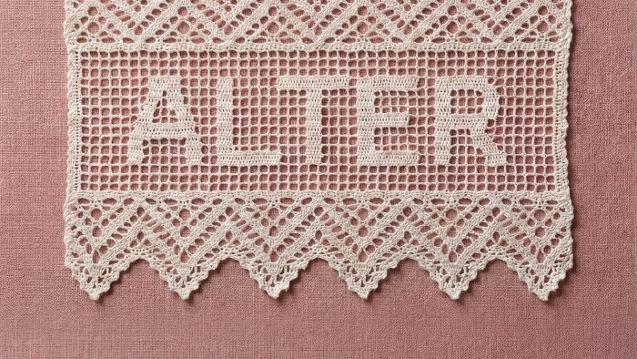 Querformatiger Detailausschnitt der H&auml;kelspitze mit dem Schriftzug &bdquo;ALTER&ldquo; auf altrosa Textilgrund. Die untere Zackenkante der Spitze ist vollst&auml;ndig sichtbar.