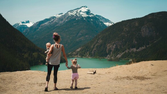 Eine Frau steht am Rande eines Sees. Auf dem Arm trägt sie ein Baby, an der anderen Hand hält sie ein kleines Mädchen. Im Hintergrund sieht man einen Berg mit schneebedecktem Gipfel.