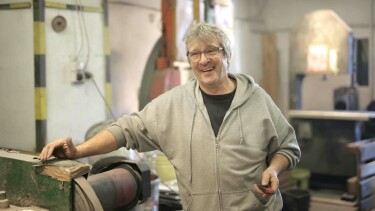 Ein alter Mann steht in einer Werkstatt an einer Maschine und lacht.
