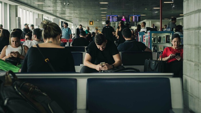 Mehrere Personen sitzen im Wartebereich am Flughafen. Alle schauen in ihre Handys und scheinen zu warten.