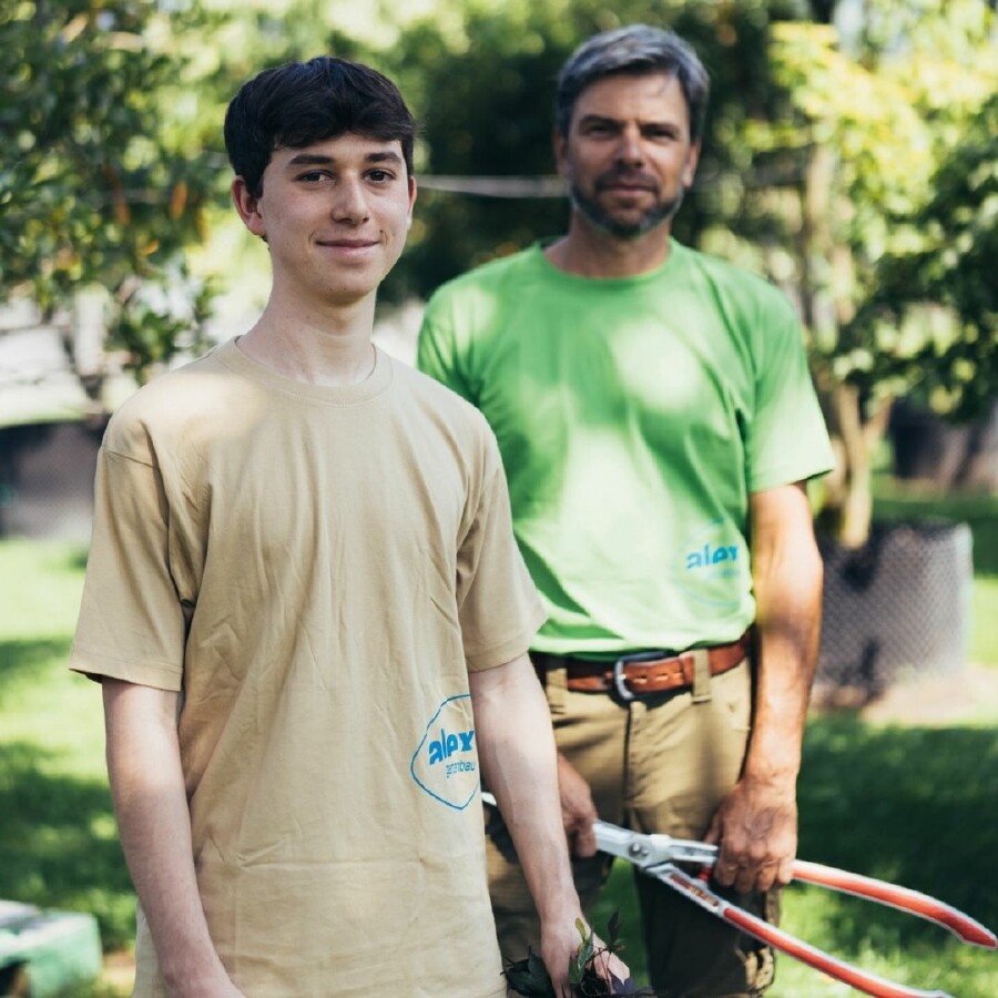 Zwei Personen im Grünen bei der Gartenarbeit: Im Vordergrund ein junger Gartenbau-Lehrling im beigen T-Shirt mit blauem Logo, im Hintergrund ein erfahrener Gärtner mit grünem Shirt und roten Gartenscheren. Symbolbild für Teamarbeit, Ausbildung im Garten- und Landschaftsbau sowie nachhaltige Berufspraxis im Freien.
