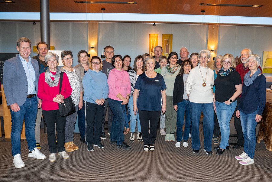Eine Gruppe von etwa 25 Personen steht eng beieinander in einem modern gestalteten Innenraum mit grauer Wandverkleidung, warmer Holzdecke und gelben Lichtakzenten. Die Teilnehmenden der Veranstaltung "Hilfe f&uuml;r die Helfenden" des letzten Turnus 2025 tragen &uuml;berwiegend Freizeitkleidung in verschiedenen Farben &ndash; darunter rote, rosa, blaue und beige Oberteile &ndash; und Jeans. Die Gruppe besteht aus Frauen und M&auml;nnern unterschiedlichen Alters, die freundlich in die Kamera blicken. Im Hintergrund sind minimalistische gelbe Wandelemente und moderne Pendelleuchten zu sehen.
