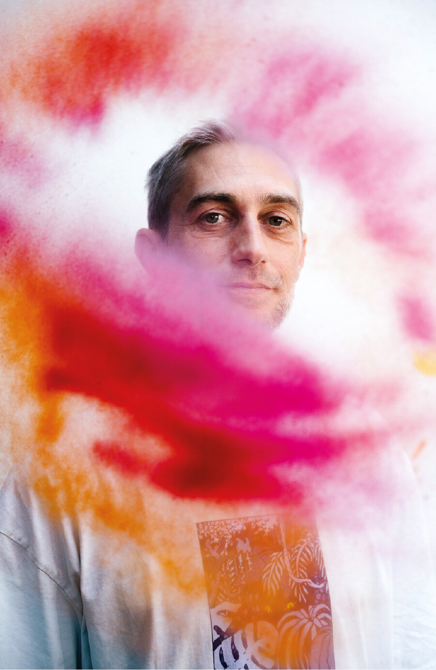 Ein Mann in einem wei&szlig;en T-Shirt spr&uuml;ht mit einer Spr&uuml;hdose leuchtend orangefarbene und rosa Farbe in die Luft. Die Farbwolke schwebt vor seinem Gesicht. Im Hintergrund ist eine helle, mit Graffiti und Farbspritzern versehene Wand zu sehen.
