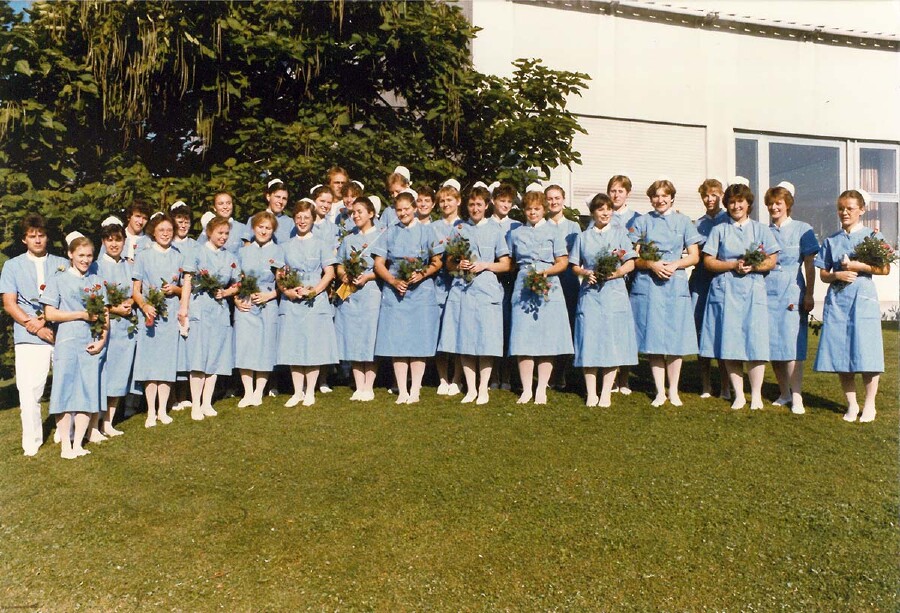 Historisches Gruppenfoto aus den 1980er-Jahren: Eine gro&szlig;e Gruppe junger Frauen in hellblauen Pflegeuniformen und wei&szlig;en Hauben steht auf einer Rasenfl&auml;che vor einem Geb&auml;ude. Die meisten halten kleine Blumenstr&auml;u&szlig;e in den H&auml;nden. Es handelt sich vermutlich um ein Abschlussfoto einer Krankenpflegeschule.