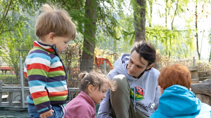 Ein Mann mit langen, braunen Haaren, Simon Engstler, kniet in einem Sandkasten. Um ihn herum stehen drei kleine Kinder, ein kleines Mädchen spielt mit ihm im Sand.