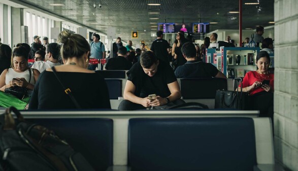 Mehrere Personen sitzen im Wartebereich am Flughafen. Alle schauen in ihre Handys und scheinen zu warten.