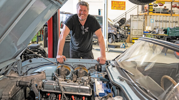 Werner Metzler l&auml;chelt in die Kamera, w&auml;hrend er sich &uuml;ber den ge&ouml;ffneten Motorraum eines Oldtimers beugt. Im Hintergrund ist eine gut ausgestattete Werkstatt zu sehen.