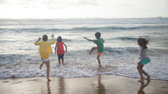 Vier Kinder spielen ausgelassen am Strand im Bereich der Brandung. Sie stehen mit dem R&uuml;cken zur Kamera und tragen bunte Sommerkleider in Gelb, Rot, Gr&uuml;n und Hellblau. Die Wellen rollen auf sie zu, eines der Kinder springt &uuml;ber das Wasser. Der Himmel ist leicht bew&ouml;lkt, das Meer rau.