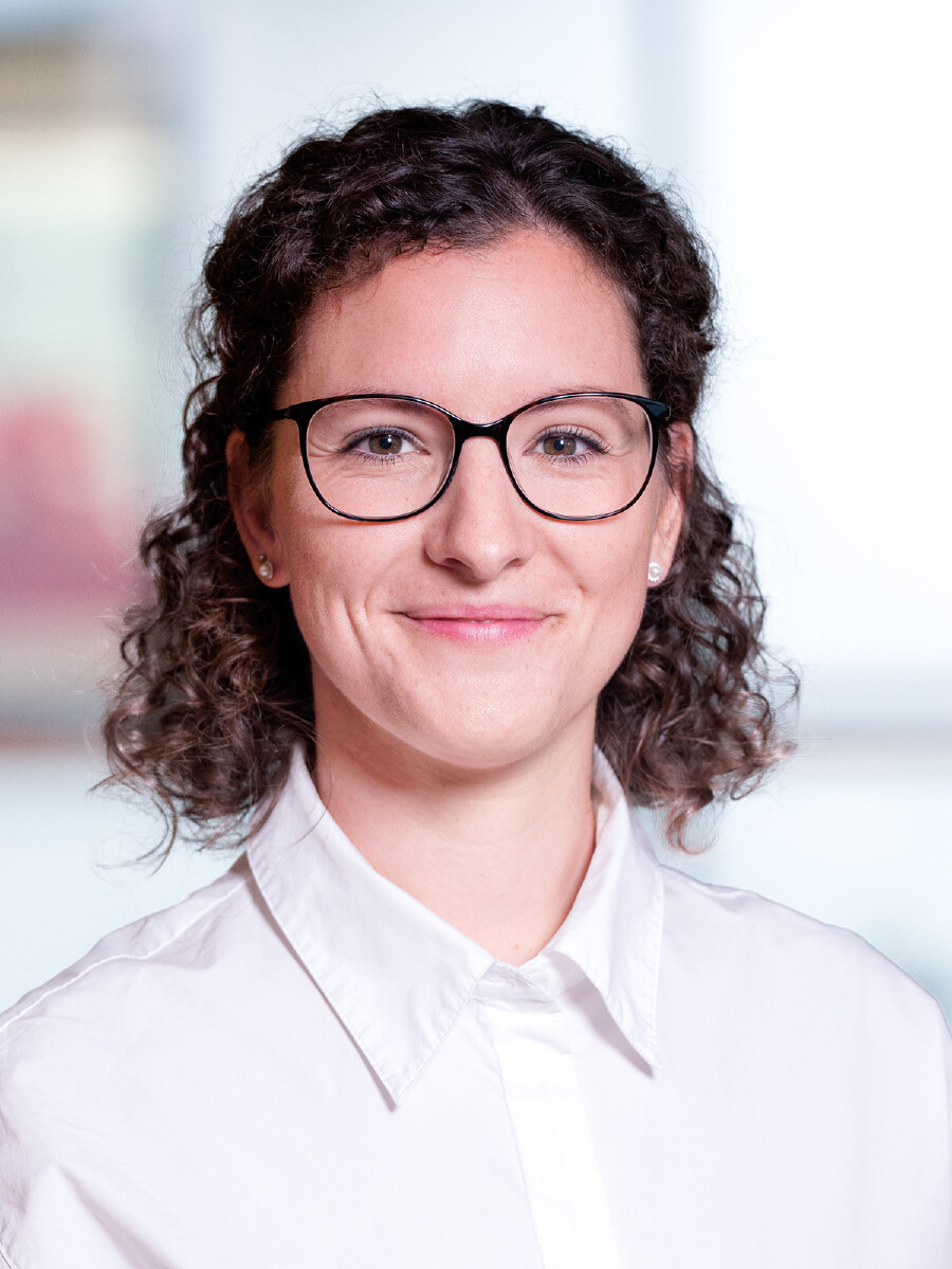 Portrait von Elena Kielwein mit braunen, lockigen haarlangen Haaren, Brille mit schwarzem Gestell und wei&szlig;em Hemd, die l&auml;chelnd in die Kamera blickt.