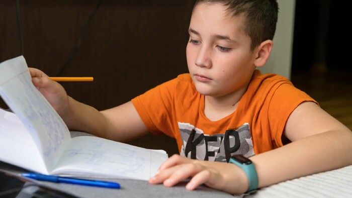 Nahaufnahme eines Jungen mit dunklen Haaren und einem orangefarbenen T-Shirt mit Aufdruck. Er sitzt an einem Tisch und blickt konzentriert in ein offenes Heft. In der rechten Hand h&auml;lt er einen gelben Stift, die linke Hand ruht auf dem Heft. Ein t&uuml;rkisfarbenes Fitnessarmband ist an seinem linken Handgelenk sichtbar. Im Vordergrund liegen ein blauer Kuli und ein Tablet. Der Hintergrund ist unscharf.