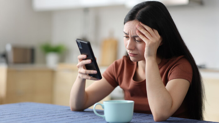 Eine Frau sitzt am Tisch und schaut verzweifelt auf ihr Smartphone.