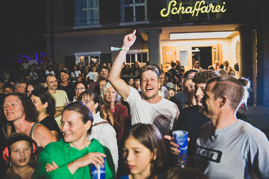 Eine Aufnahme einer jubelnden Menschenmenge bei Nacht vor dem beleuchteten &bdquo;Schaffarei&ldquo;-Geb&auml;ude. Ein junger Mann im Mittelpunkt hebt lachend den Arm und den Finger hoch, umgeben von anderen fr&ouml;hlichen Festivalbesuchern.